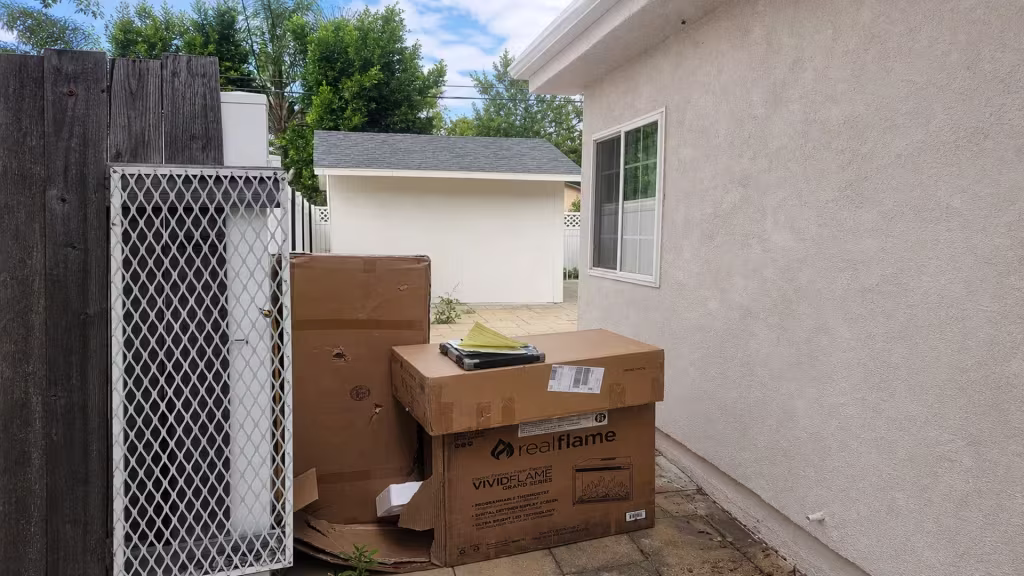 Before installation, a narrow side yard is blocked by a weathered wood fence, metal mesh panel, and stacked cardboard boxes.
