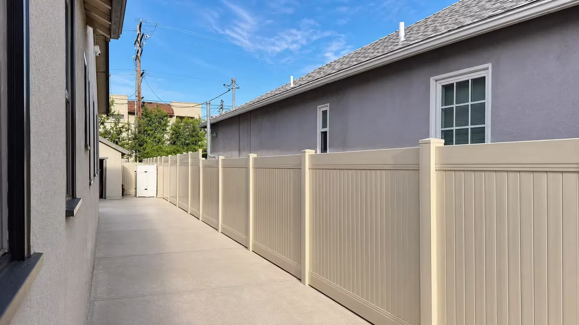 Roof vents and pipes overlook this Tan vinyl privacy fence lining a cracked concrete passage in Glendale