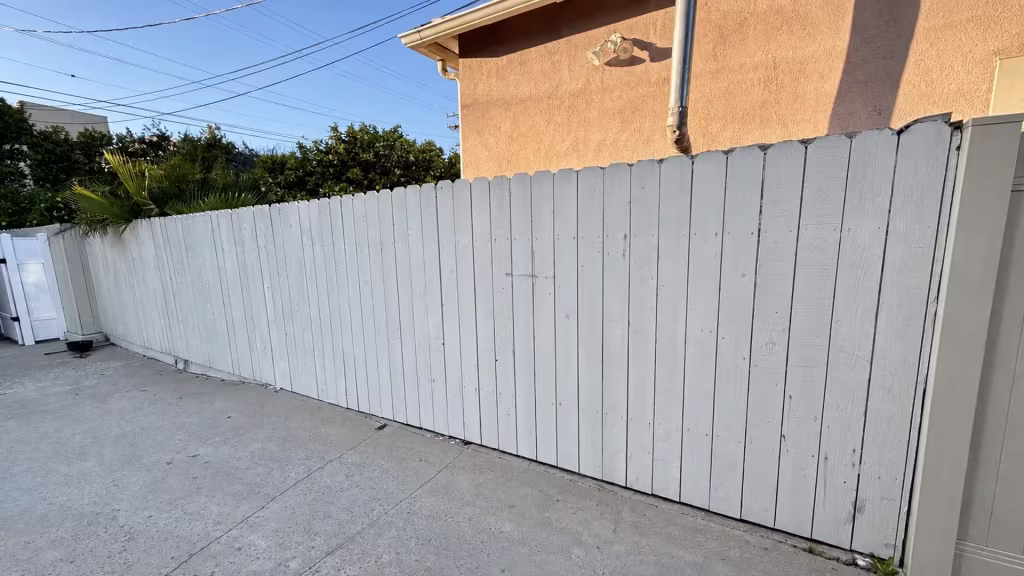 Before installation, a long white-painted wood fence borders a concrete patio beside a tan stucco house.