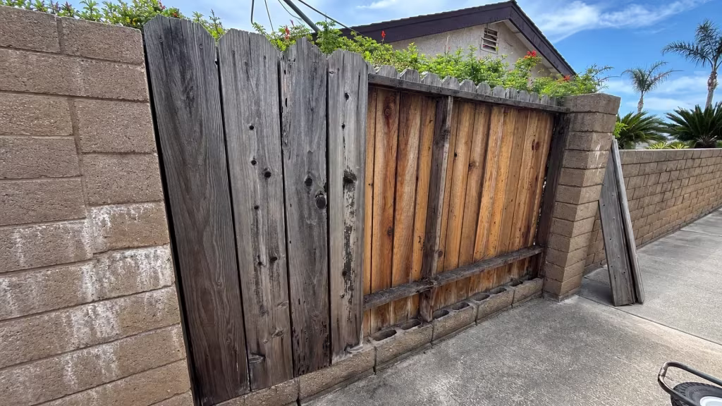 Before installation, a weathered mismatched wood fence and gate stand between tan block columns along a concrete driveway.
