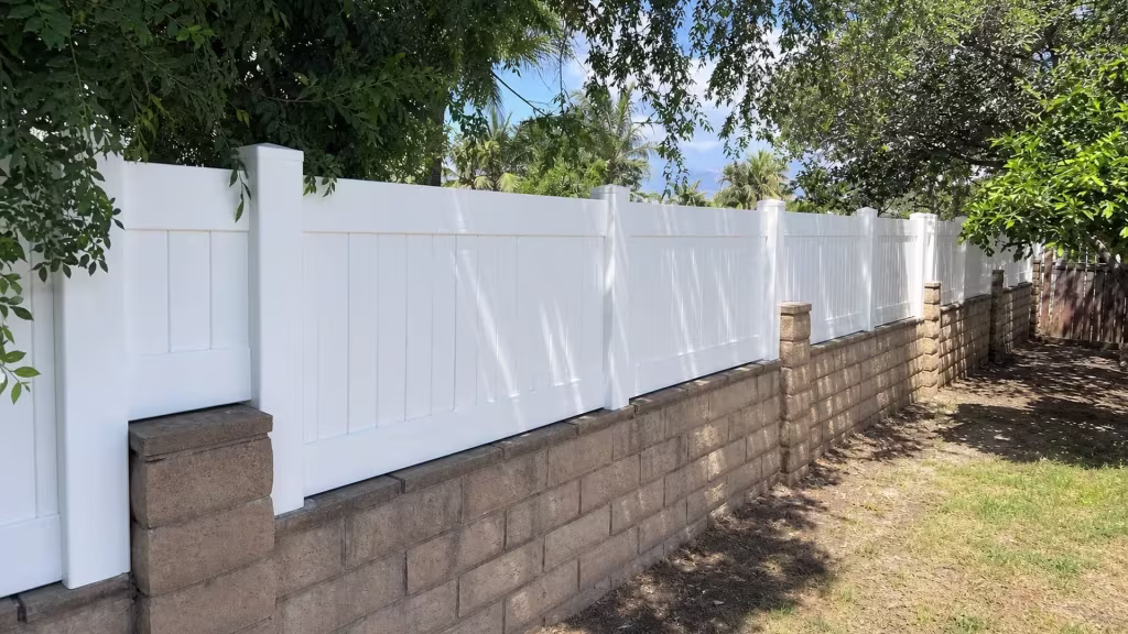 Broadleaf trees and palms overhang a White vinyl topper that lines this Azusa backyard wall for privacy