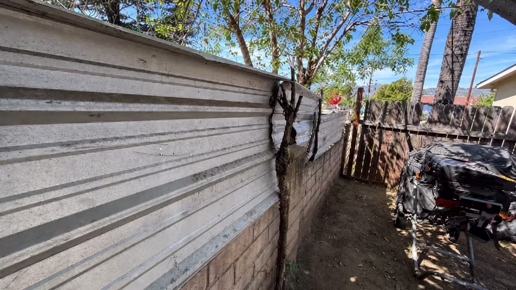 Before installation, a damaged corrugated metal barrier, old wood gate, tree trunk, and covered equipment fill a dirt side yard.