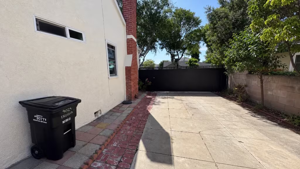 Before installation, a driveway beside the house ends at a black metal gate near a trash bin, pavers, and trees.