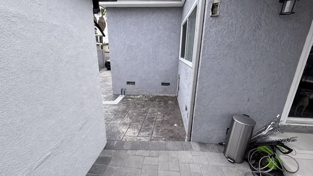 Before installation, an open paved passage runs between gray stucco walls with a trash can and coiled hoses nearby.