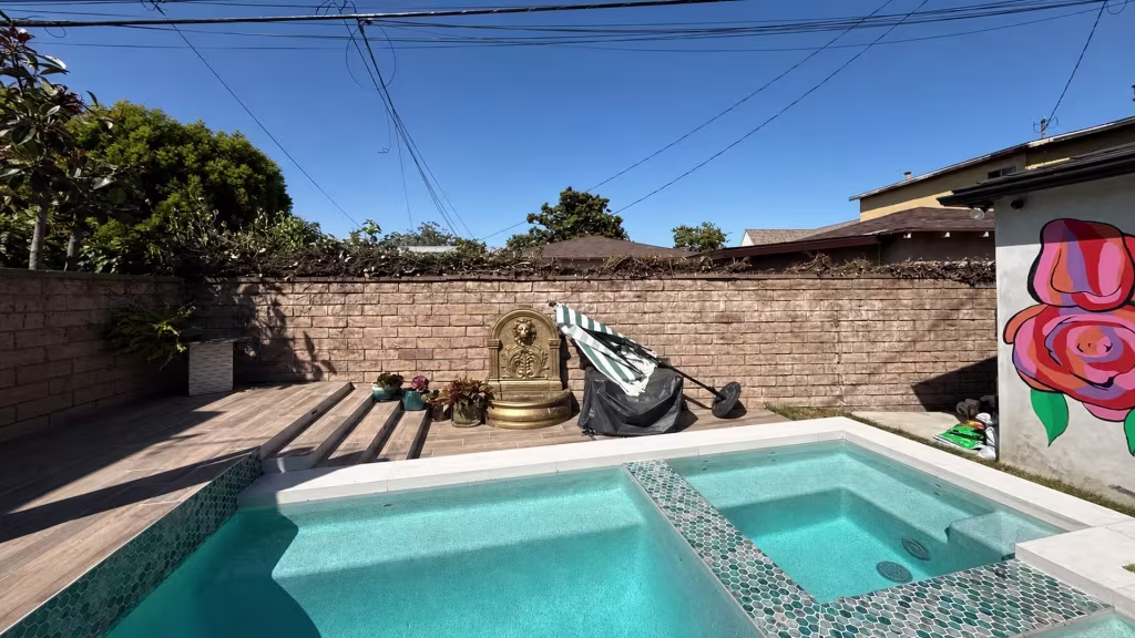 Before installation, a backyard pool sits beside a bare brick wall with trimmed branches, a fountain, and a painted mural.