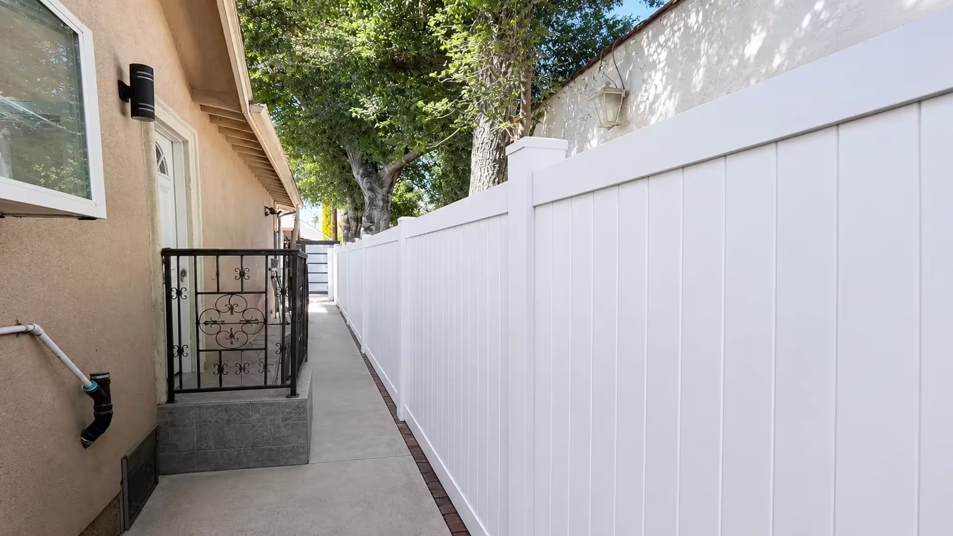 Brick edging borders the narrow concrete walkway lined by this White vinyl privacy fence in Pasadena