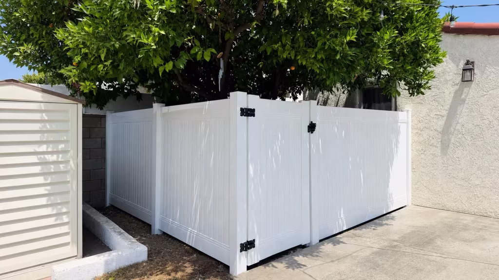 A mature citrus tree and a hanging wind chime stand beside this White vinyl privacy wall topper near a beige shed