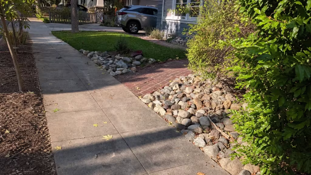 Before installation, an open front lawn and walkway feature rock drainage beds and dense shrubs without a boundary fence.