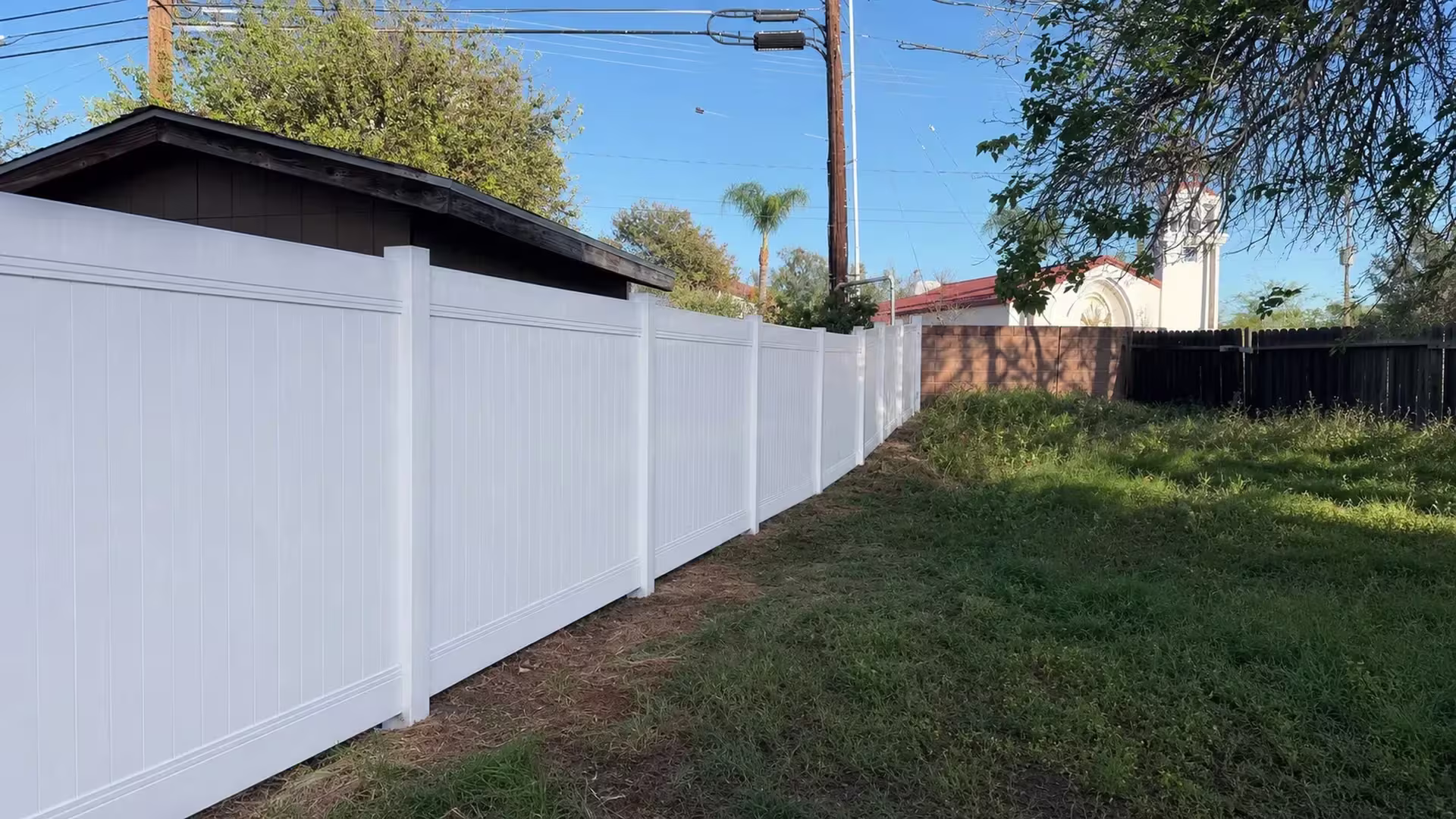 A cream-colored building with a red tile roof rises beyond the line where this vinyl privacy fence runs through Orange