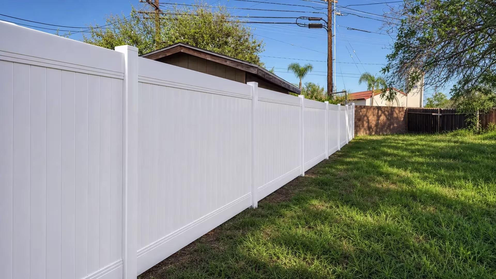 An uneven backyard lawn runs beneath this White vinyl privacy fence beside a brown masonry wall in North Hollywood