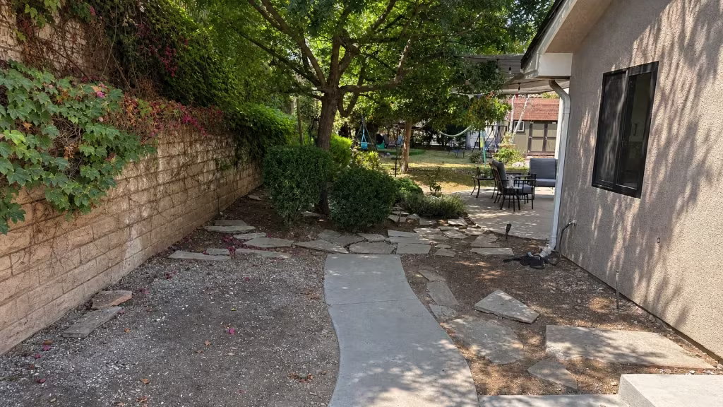 Before installation, a shaded side yard has bare soil, stepping stones, an ivy-covered wall, and patio furniture.