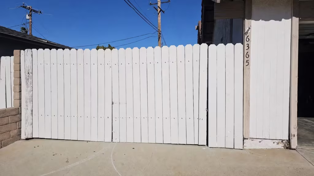 Before installation, a white wood fence and gate close off a driveway beside the garage.