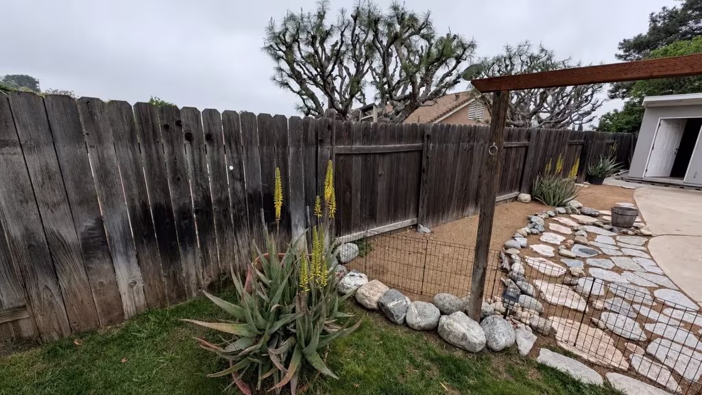 Before installation, a weathered wood fence and wire garden barrier surround a sandy yard with stepping stones and aloe.