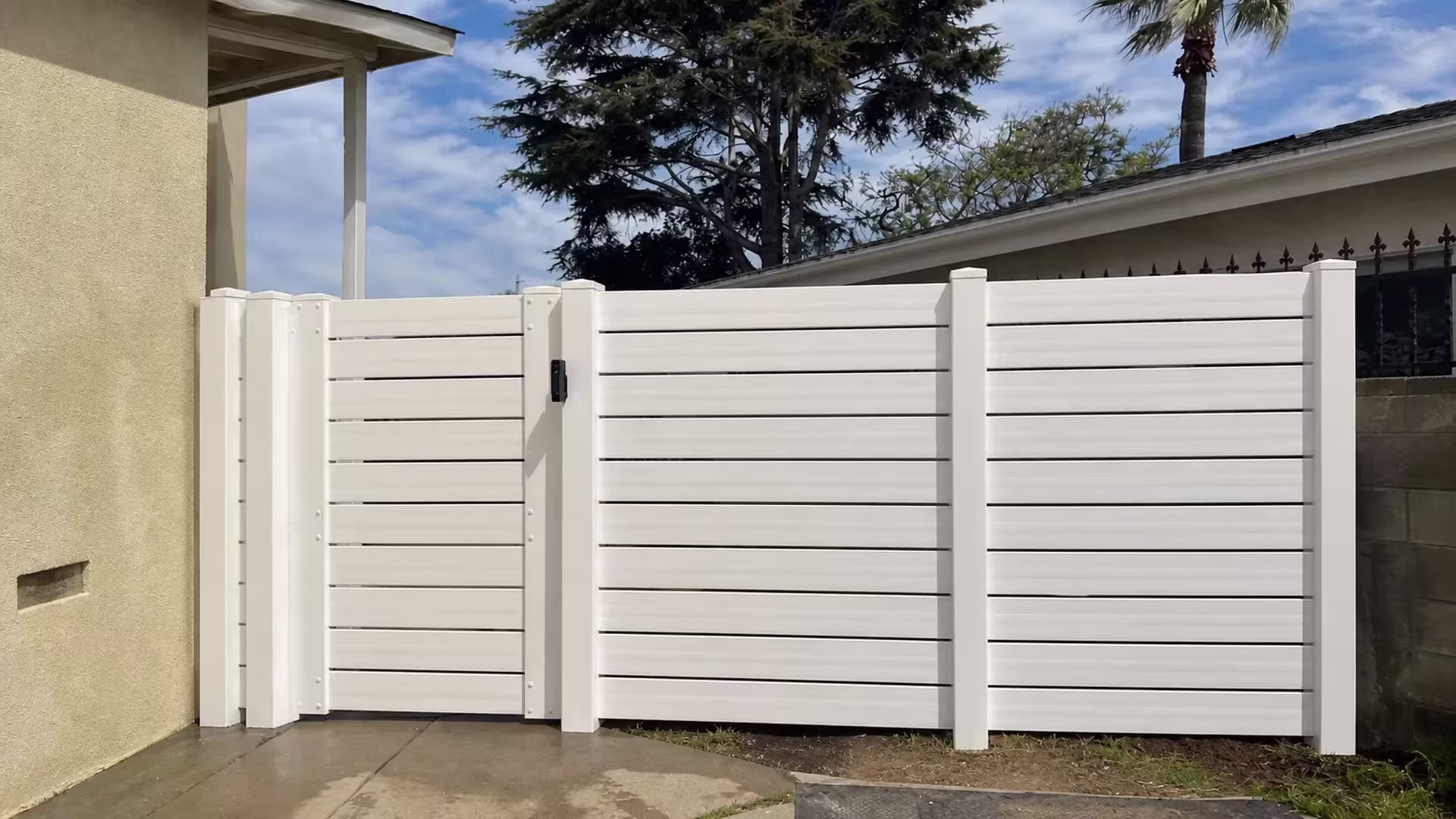 White vinyl horizontal picket fence and gate front a pale stucco wall, with a black spear-top metal fence visible behind them