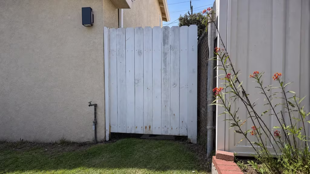 Before installation, a white wood gate stands between a stucco house and chain-link boundary beside a lawn and flowers.