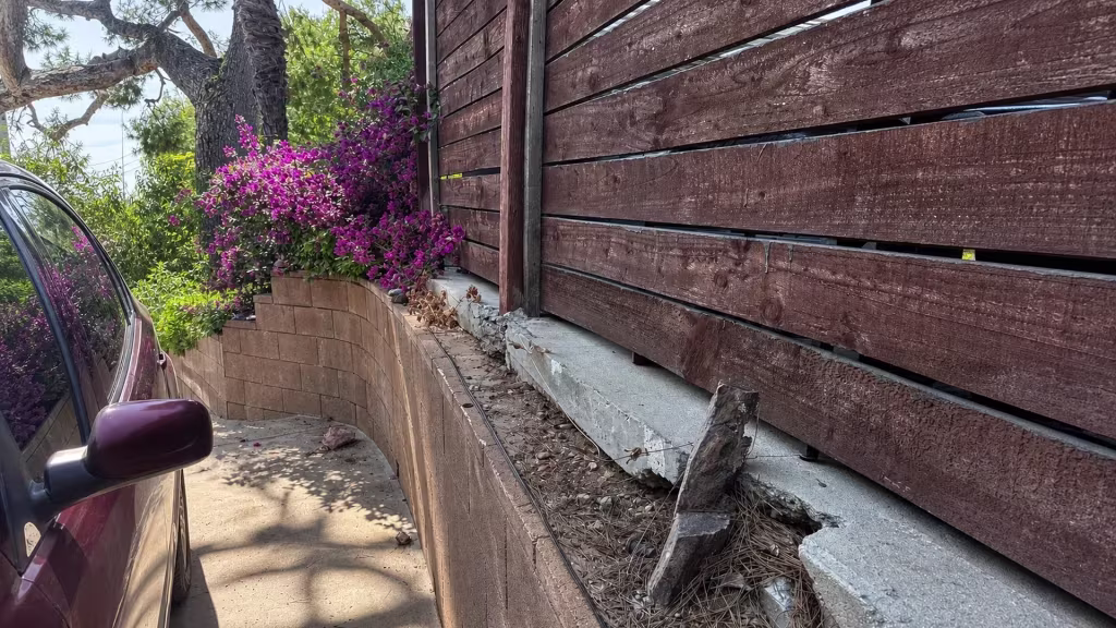 Before installation, a dark horizontal wood fence sits on a damaged concrete edge beside a narrow driveway and flowering shrubs.
