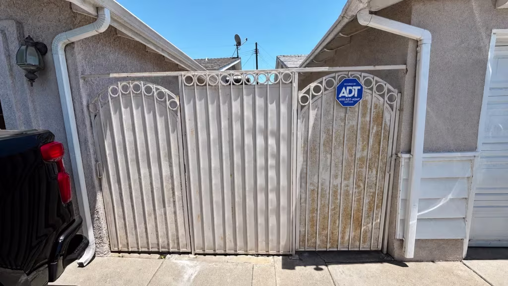 Before installation, rusted white metal double gates span a concrete driveway between two stucco buildings.
