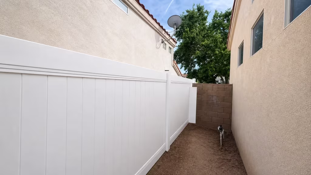 White vinyl privacy fence ends near a cracked stucco wall and satellite dish in a Palmdale passage