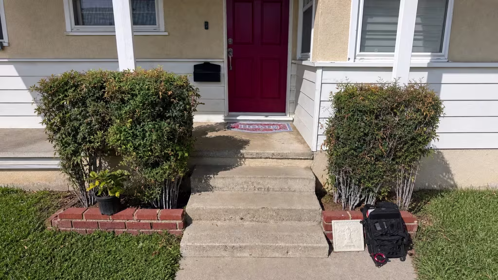 Before installation, the front porch has a red door, concrete steps, and trimmed shrubs with no fence present.
