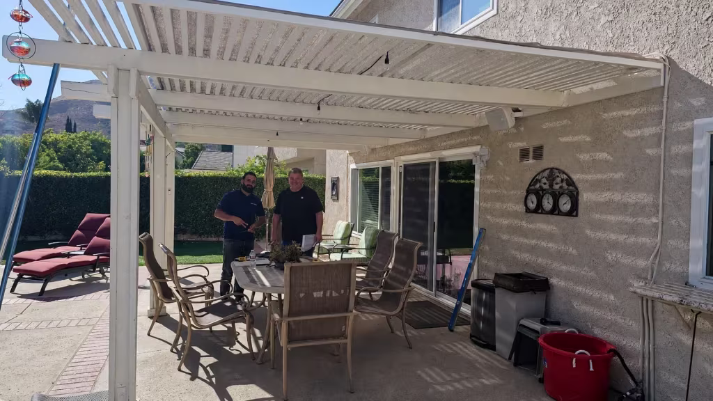 Before installation, an older white slatted patio cover extends over an outdoor dining area beside the house.