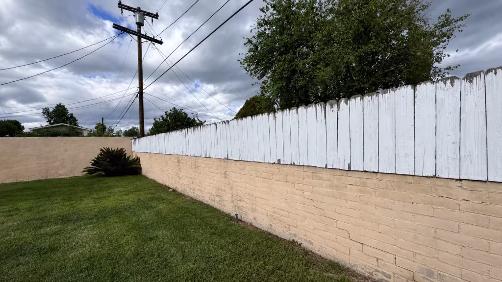 Before installation, worn white-painted wood boards top a tan brick wall along the lawn.
