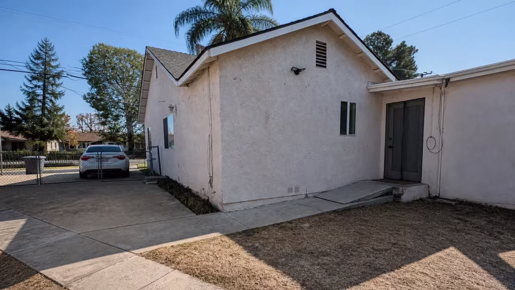 Before installation, an open concrete side yard beside a stucco house ends at a chain-link driveway gate.