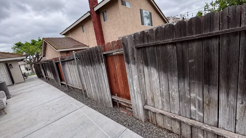Before installation, a weathered mismatched wood fence with several leaning panels borders a paved side yard.