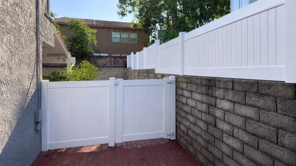 Between a stucco house wall and a block retaining wall, a matching gate completes this White vinyl privacy wall topper's run