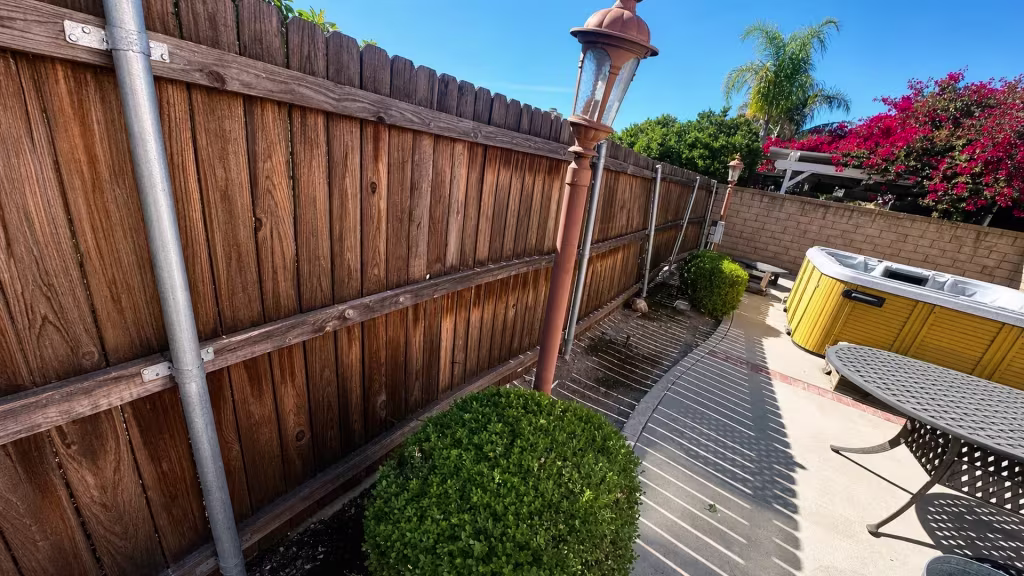 Before installation, a tall wood fence reinforced with metal posts borders a patio and hot tub.