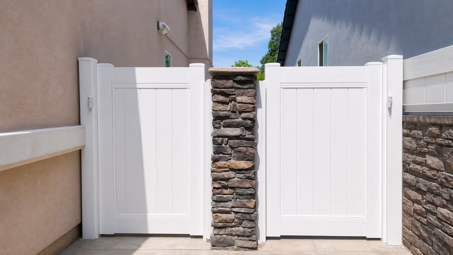 A stacked-stone masonry pier separates gate leaves belonging to a White vinyl fence between stucco homes in Duarte
