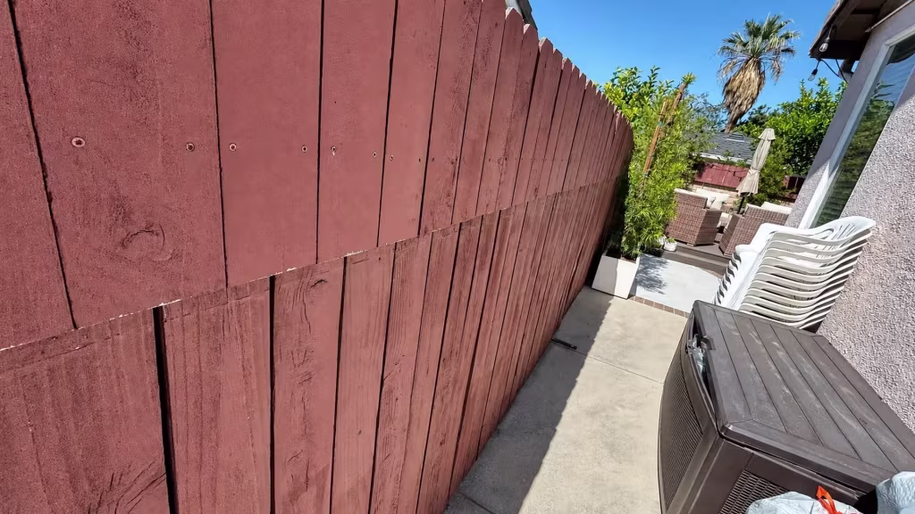 Before installation, a red-painted wood fence borders a narrow concrete patio with stacked chairs and a storage bin.