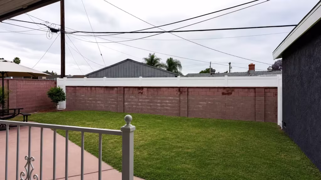 A scrolled porch railing with a round finial rises before this White vinyl privacy fence topping a wall in West Covina