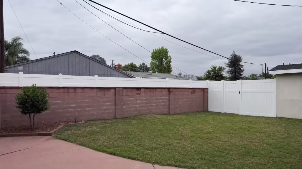 A cracked concrete walkway edge runs beside this White vinyl fence near the reddish brown block wall in West Covina