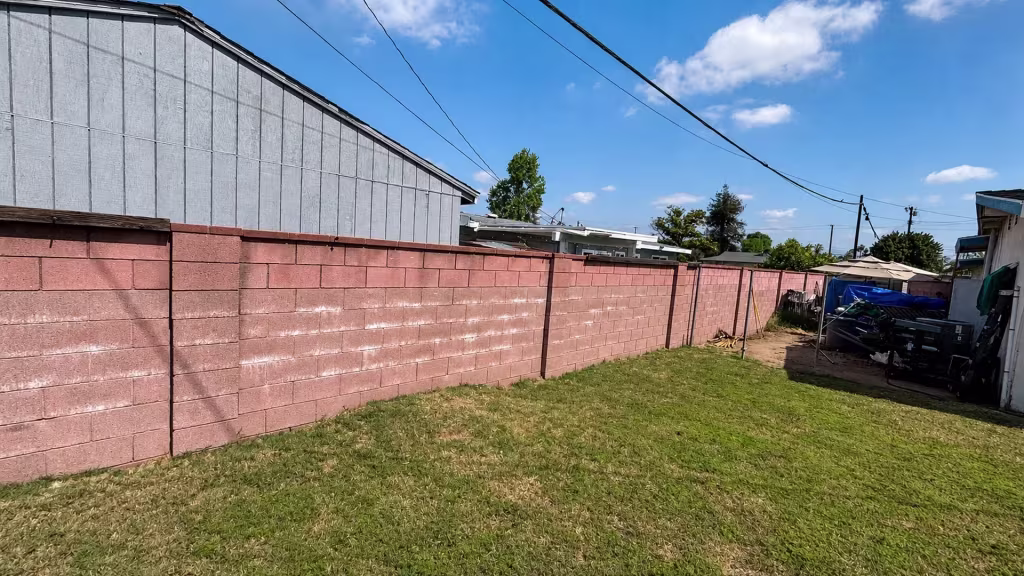 Before installation, a long pink block wall borders a grassy yard with stored items at the far end.