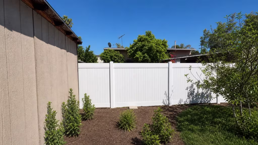 A tan outbuilding and a large branching tree frame this White vinyl privacy fence across the rear yard in North Hollywood