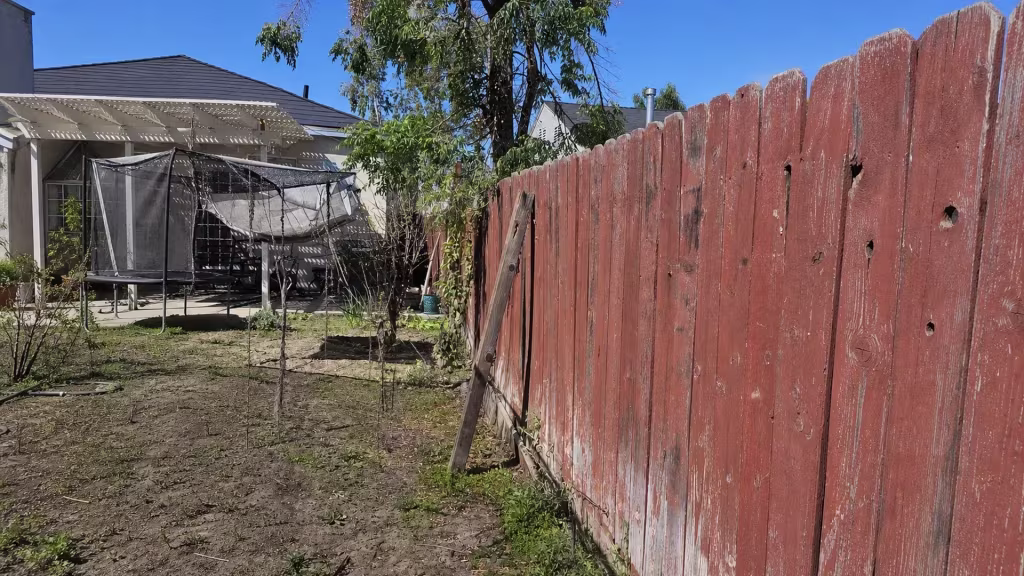 Before installation, a faded red wood fence borders a bare garden area and trampoline.
