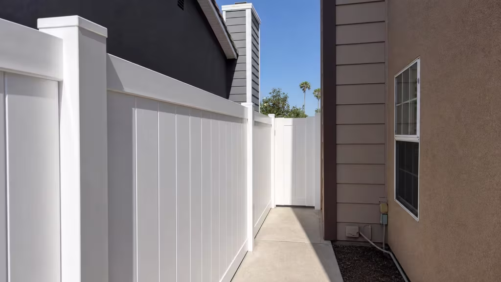 White vinyl vertical picket fence lines a narrow concrete side passage between two Chino houses