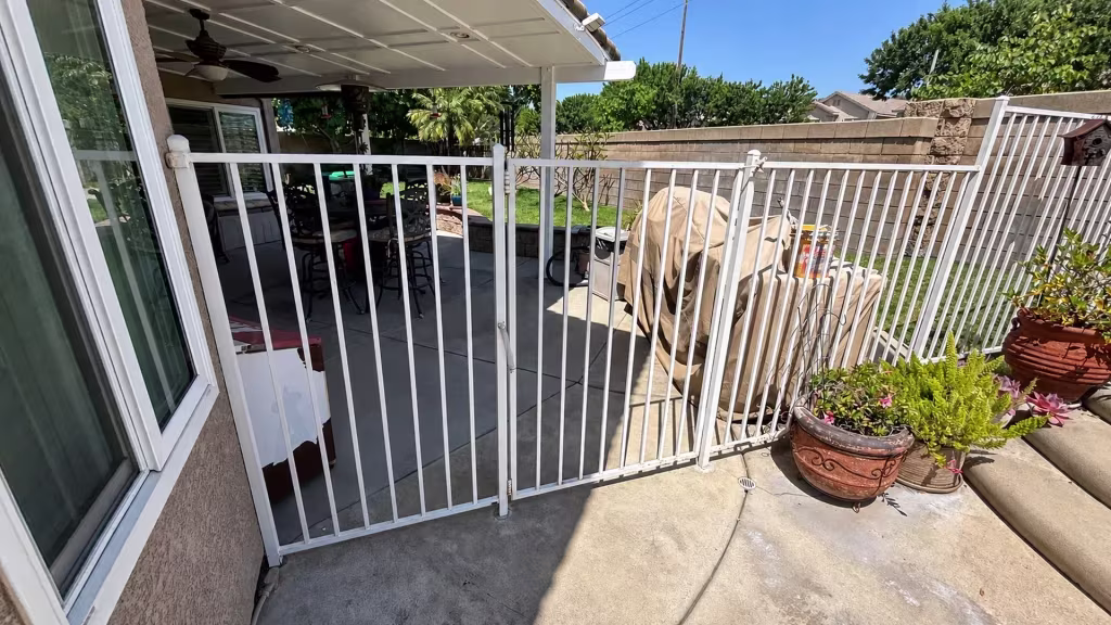 Before installation, a white metal safety gate encloses a covered patio with a grill and potted plants.