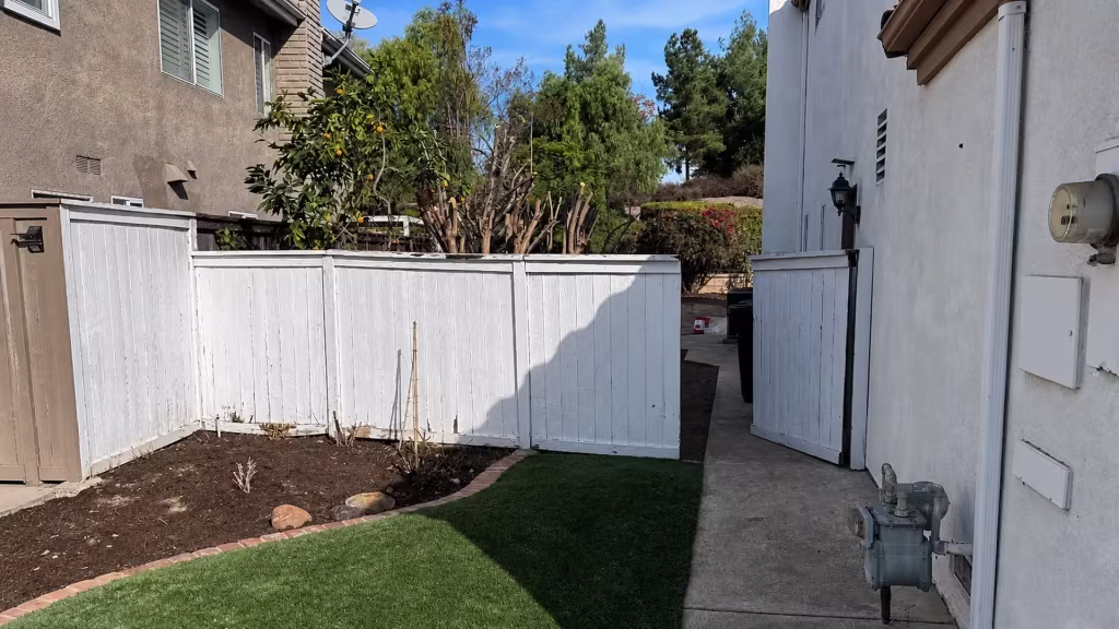 Before installation, a white wood fence and open gate enclose a lawn and garden between two houses.