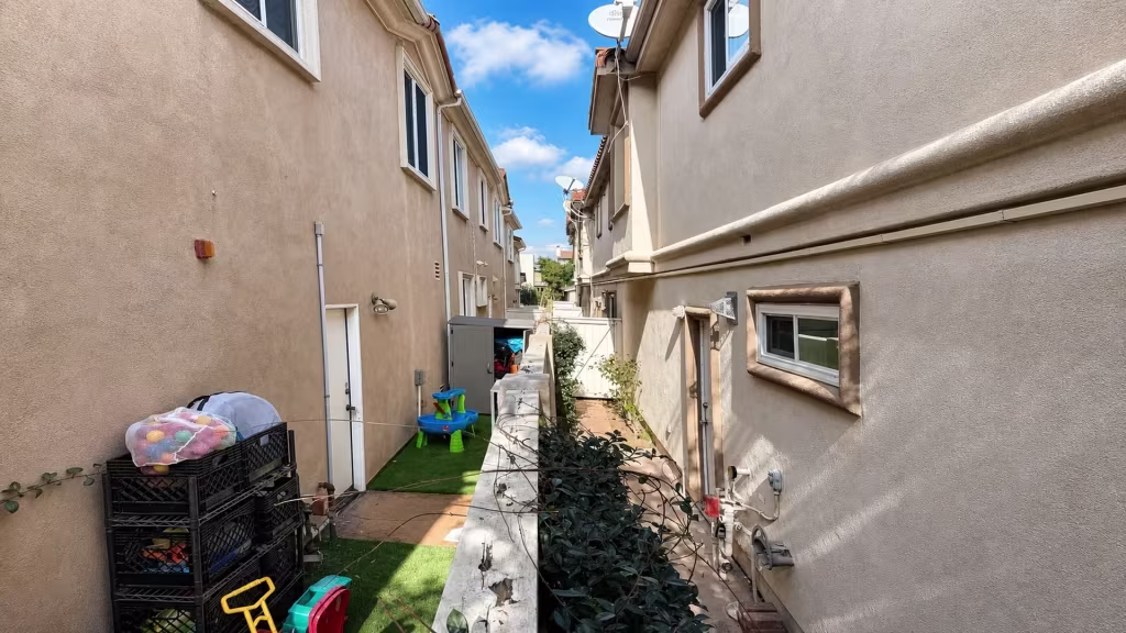 Before installation, narrow shared side yards between stucco buildings contain a low wall, artificial grass, toys, and stored items.