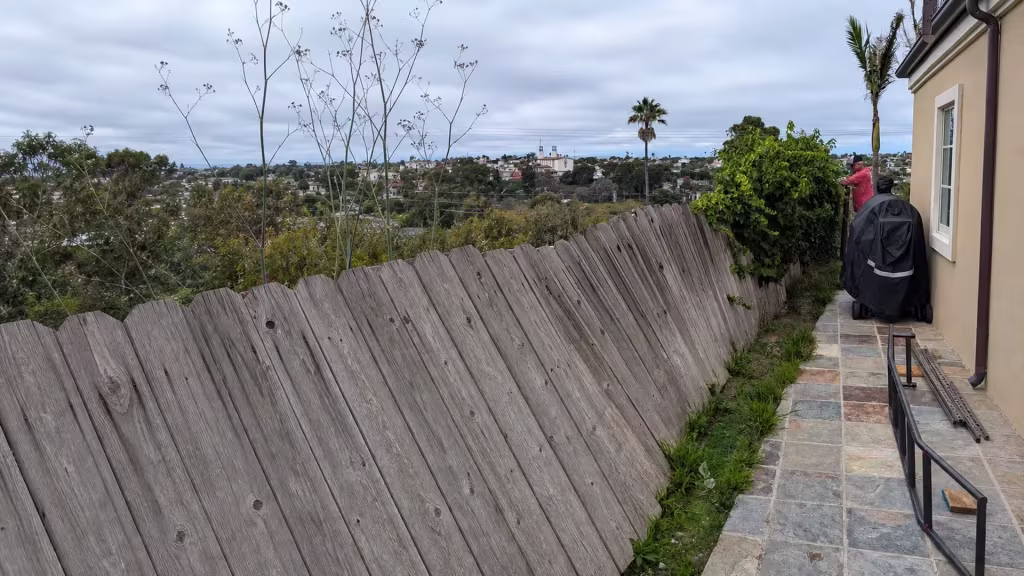 Before installation, a severely leaning gray wood fence borders a tiled side patio overlooking a hillside neighborhood.