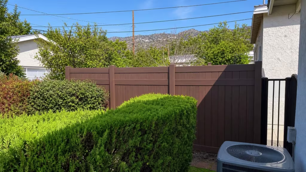 An outdoor AC condenser sits near this Dark Sequoia vinyl privacy fence crossing the backyard below a hillside in Burbank