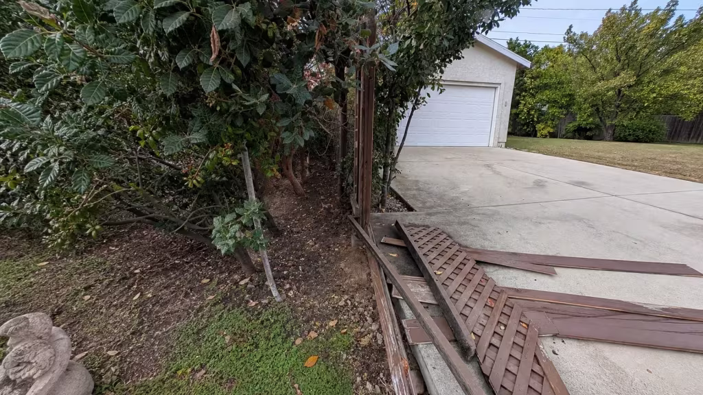 Before installation, dismantled wood lattice and boards are piled beside a driveway and garage near dense shrubs.