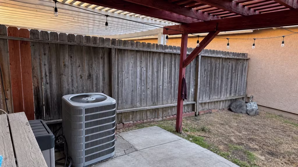 Before installation, a weathered wood fence stands beneath a pergola and string lights beside an air-conditioning unit and bare soil.