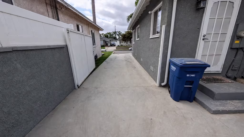 Before installation, an open white driveway gate stands beside a gray stucco house and blue trash bin.