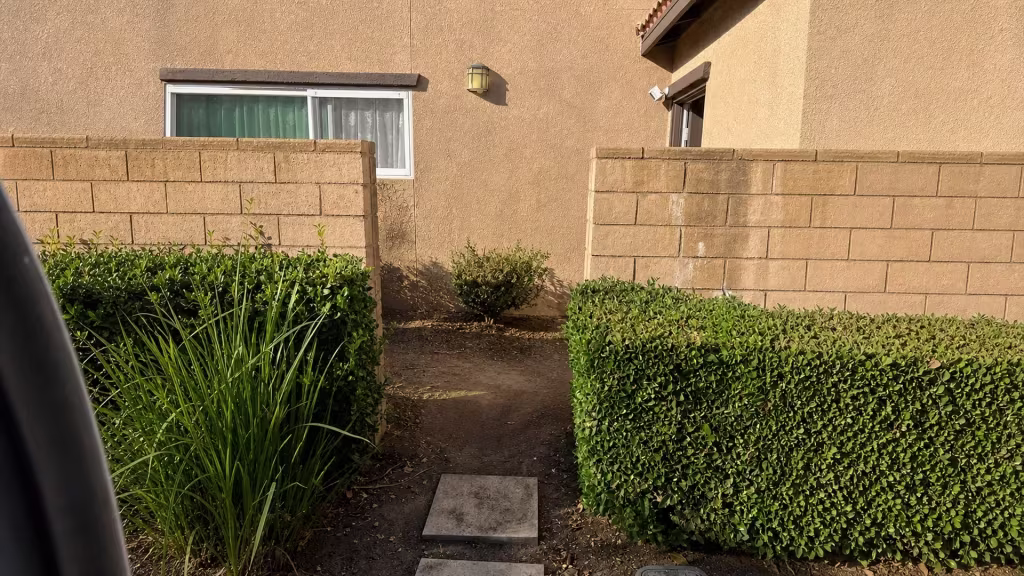 Before installation, a dirt path and trimmed hedges fill the gap between two tan block walls.