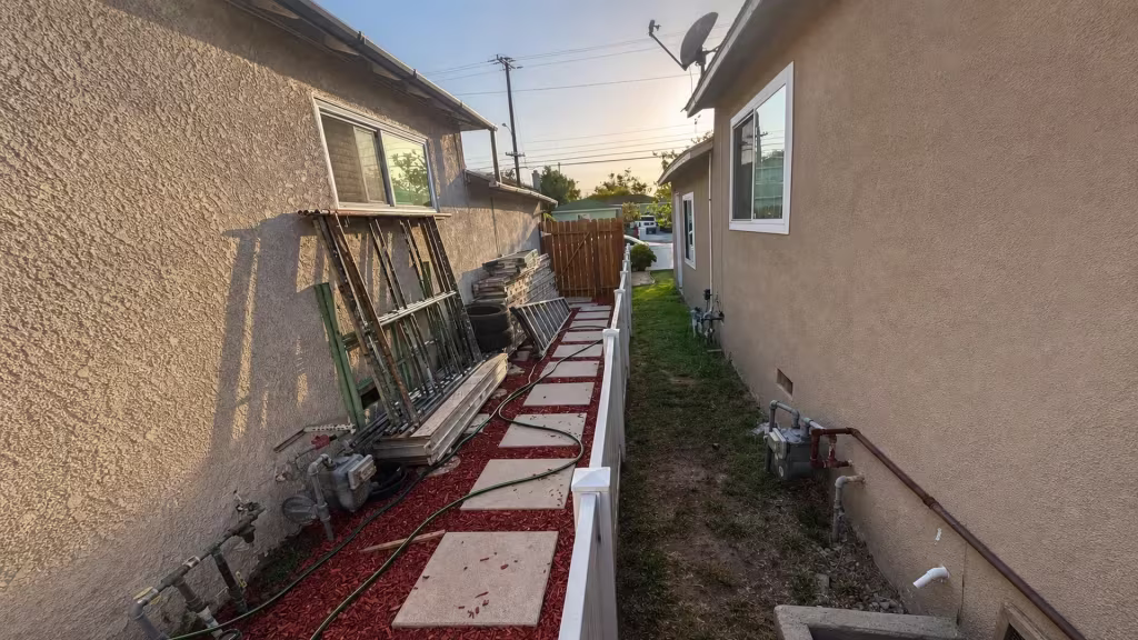 Before installation, a narrow passage between houses contains a low white divider, stored ladders, materials, and stepping stones.