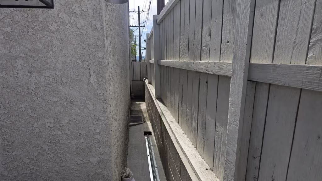 Before installation, a very narrow concrete passage runs between a stucco wall and tall gray-painted wood fence.