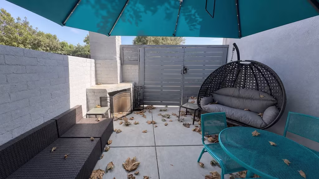 Before installation, a shaded patio has a gray horizontal-slat metal gate, white brick fireplace, hanging chair, and outdoor furniture.