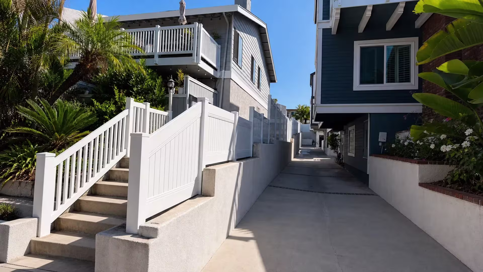 A dark blue-gray house with a bay window faces this White vertical vinyl picket fence along a Redondo Beach driveway